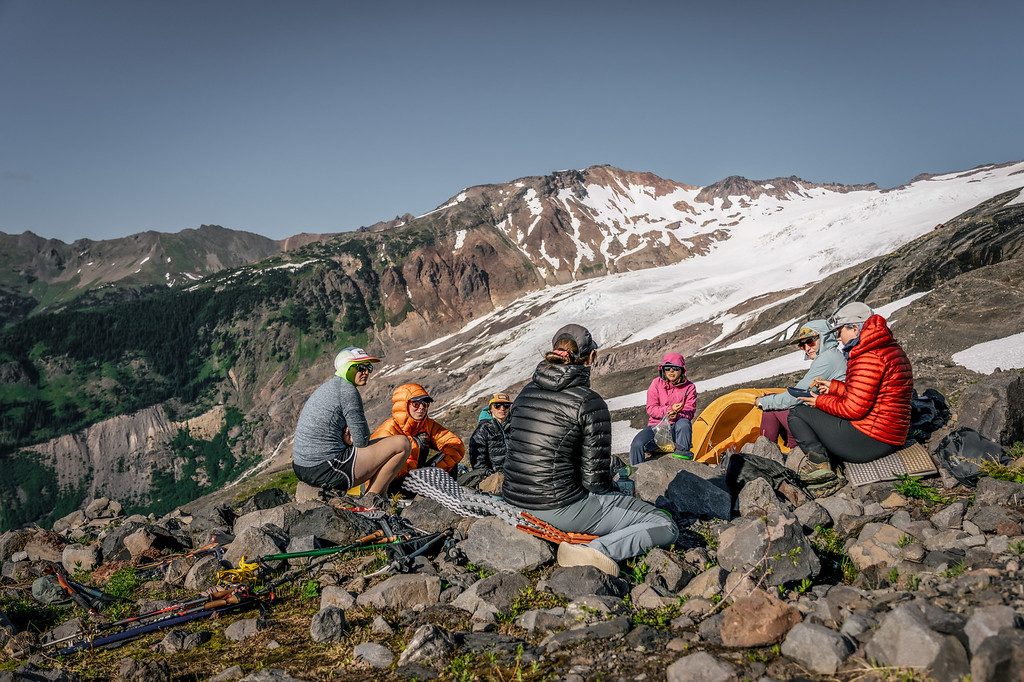 A group of women in down jackets sitting in a circle at a glacier camp beneath a snowy ridge.
