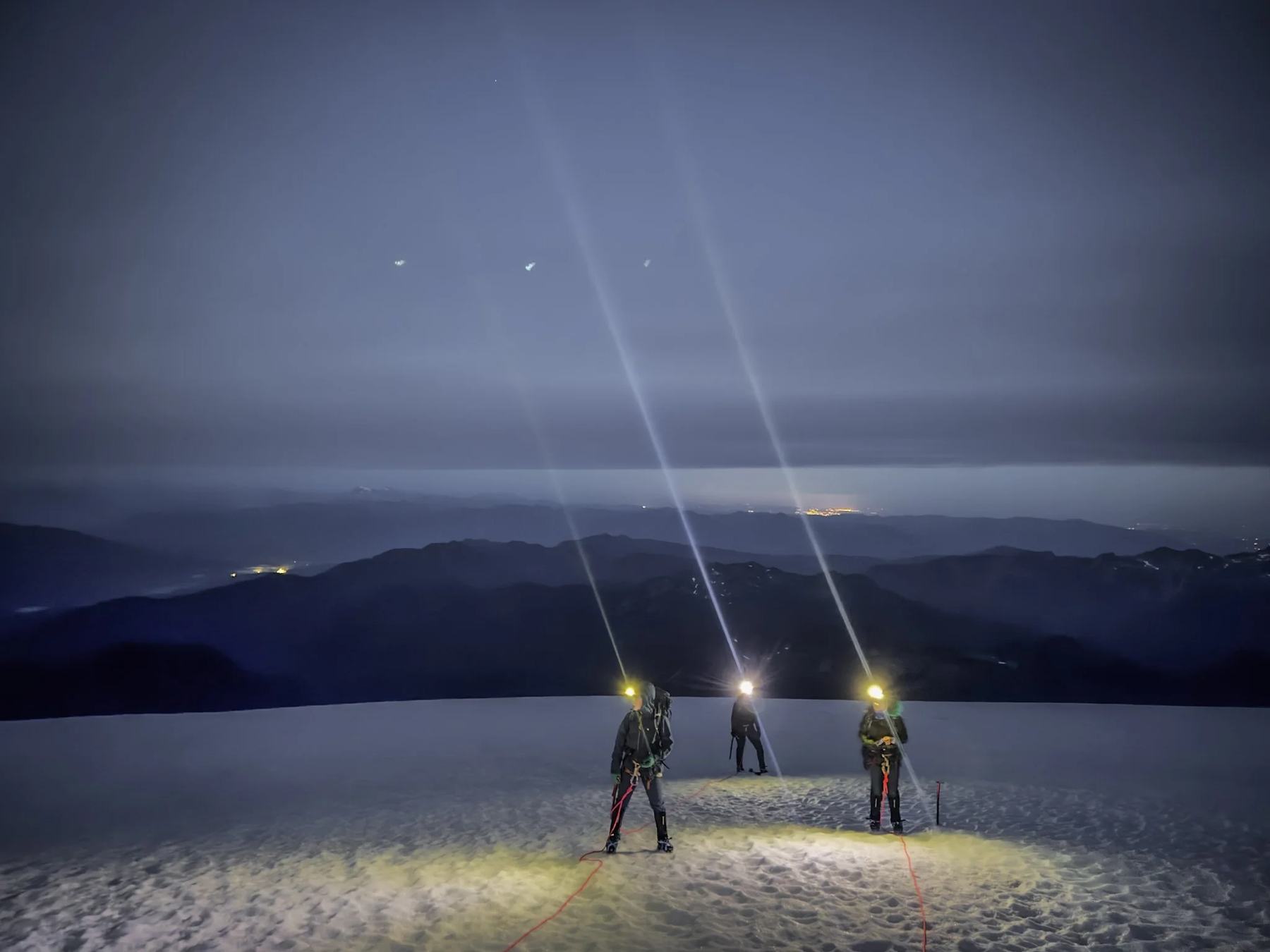 Three climbers roped together on a glacier at night, headlamp beams pointing at the sky.